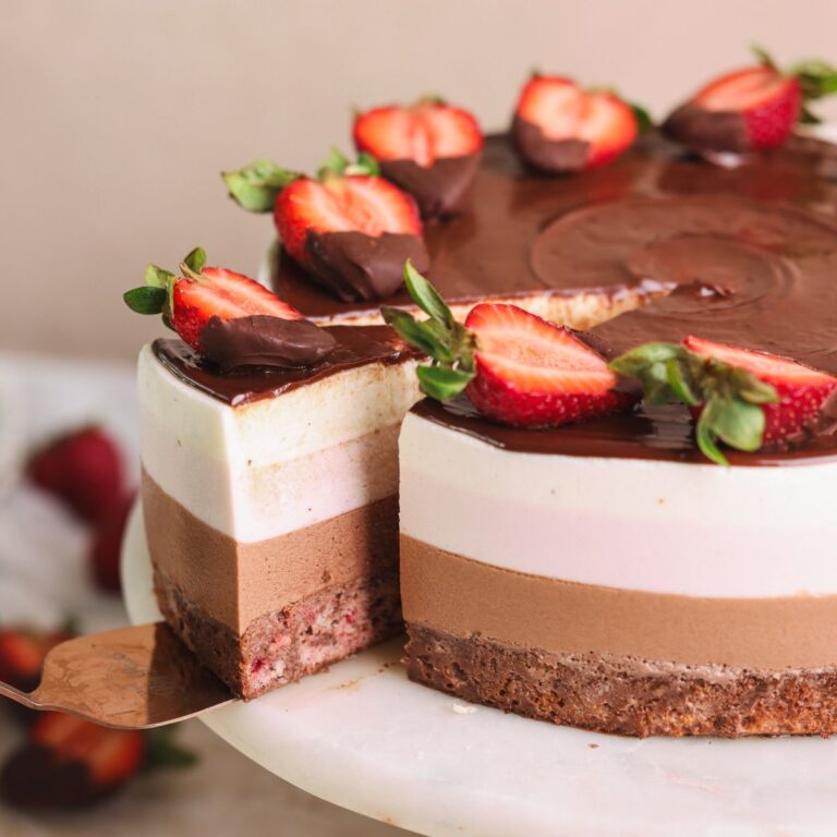 neopolitan ice cream mousse cake on a cake stand with a spatula pulling out a slice. the cake has layers of strawberry cake, chocolate mousse, strawberry mousse and vanilla mousse, and is decorated with ganache and strawberries