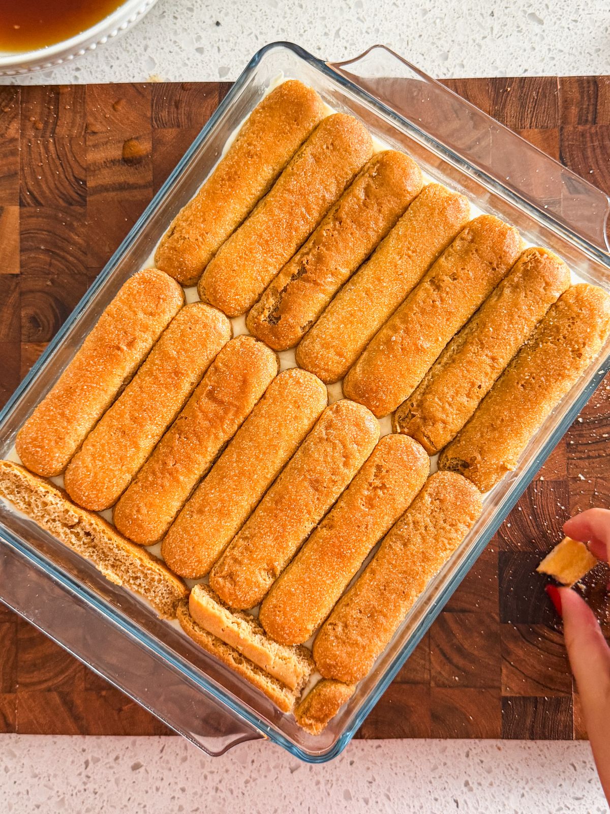 A glass dish filled with neatly arranged chai soaked ladyfinger cookies on a wooden board. A hand dips a ladyfinger in chai, preparing it to add to the dish.