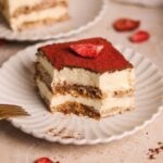 A slice of chai tiramisu topped with a dusting of cocoa powder and a crushed freeze dried strawberries sits on a white scalloped plate, with a fork and another dessert slice in the background.