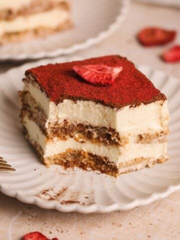 A slice of chai tiramisu topped with a dusting of cocoa powder and a crushed freeze dried strawberries sits on a white scalloped plate, with a fork and another dessert slice in the background.