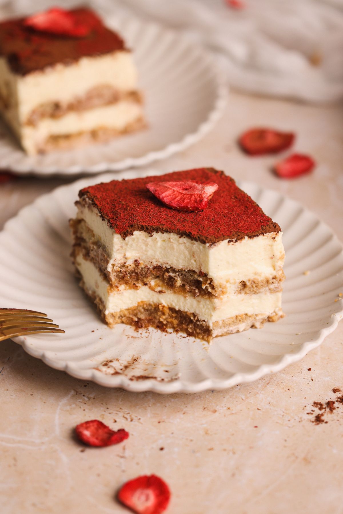 A slice of chai tiramisu topped with cocoa powder and freeze dried strawberries sits on a white scalloped plate. Another slice is visible in the background, with strawberry pieces scattered on the table.