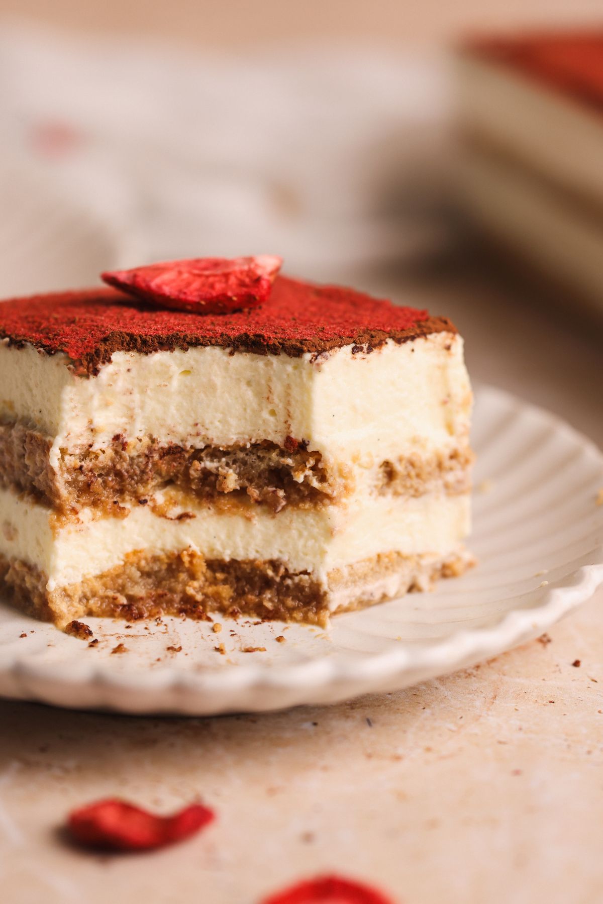 A slice of chai tiramisu with layers of cream and chai soaked ladyfingers sits on a white plate, topped with cocoa powder and a freeze dried strawberries. The background is softly blurred.