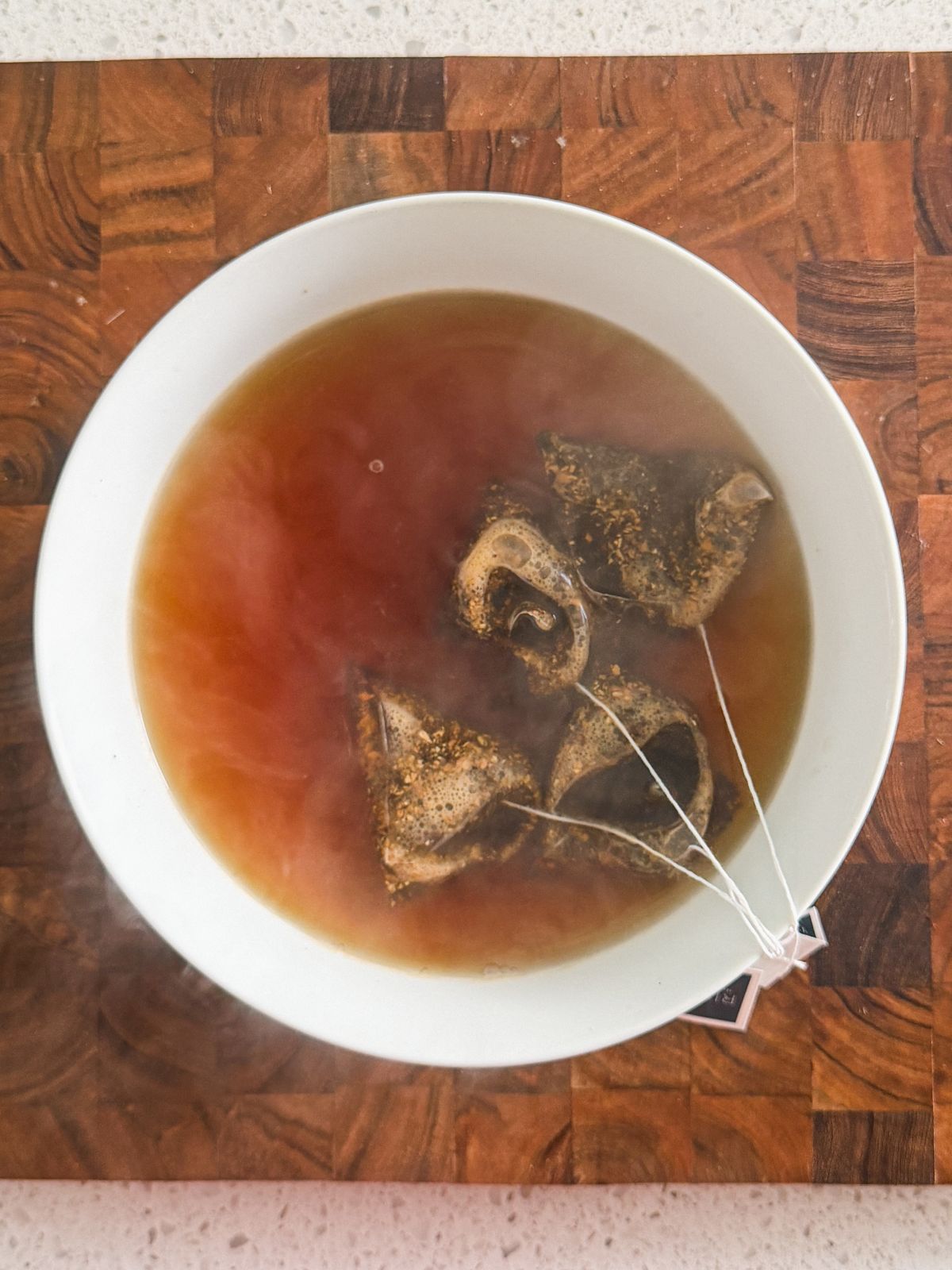 A white bowl filled with hot tea, with four steeping tea bags floating on the surface. The bowl is placed on a wooden surface with a checkered pattern. Steam rises from the tea.