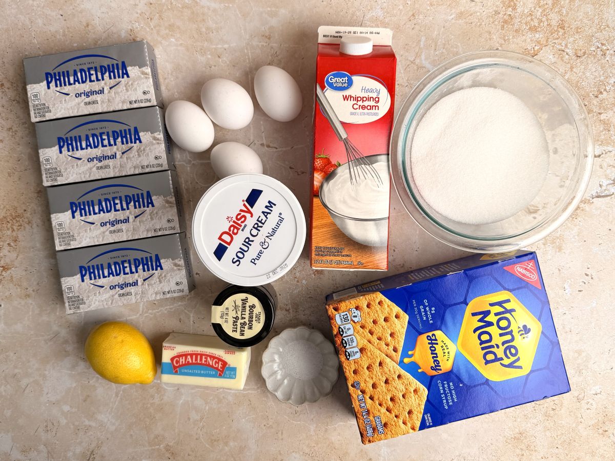 Blocks of cream cheese, four eggs, heavy whipping cream, a bowl of sugar, sour cream, graham crackers, a lemon, a stick of butter, and a small dish of salt are arranged on a countertop.