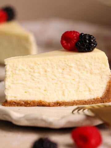 A slice of creamy cheesecake with a graham cracker crust, topped with a fresh raspberry and blackberry, is served on a ceramic plate with a gold fork nearby.