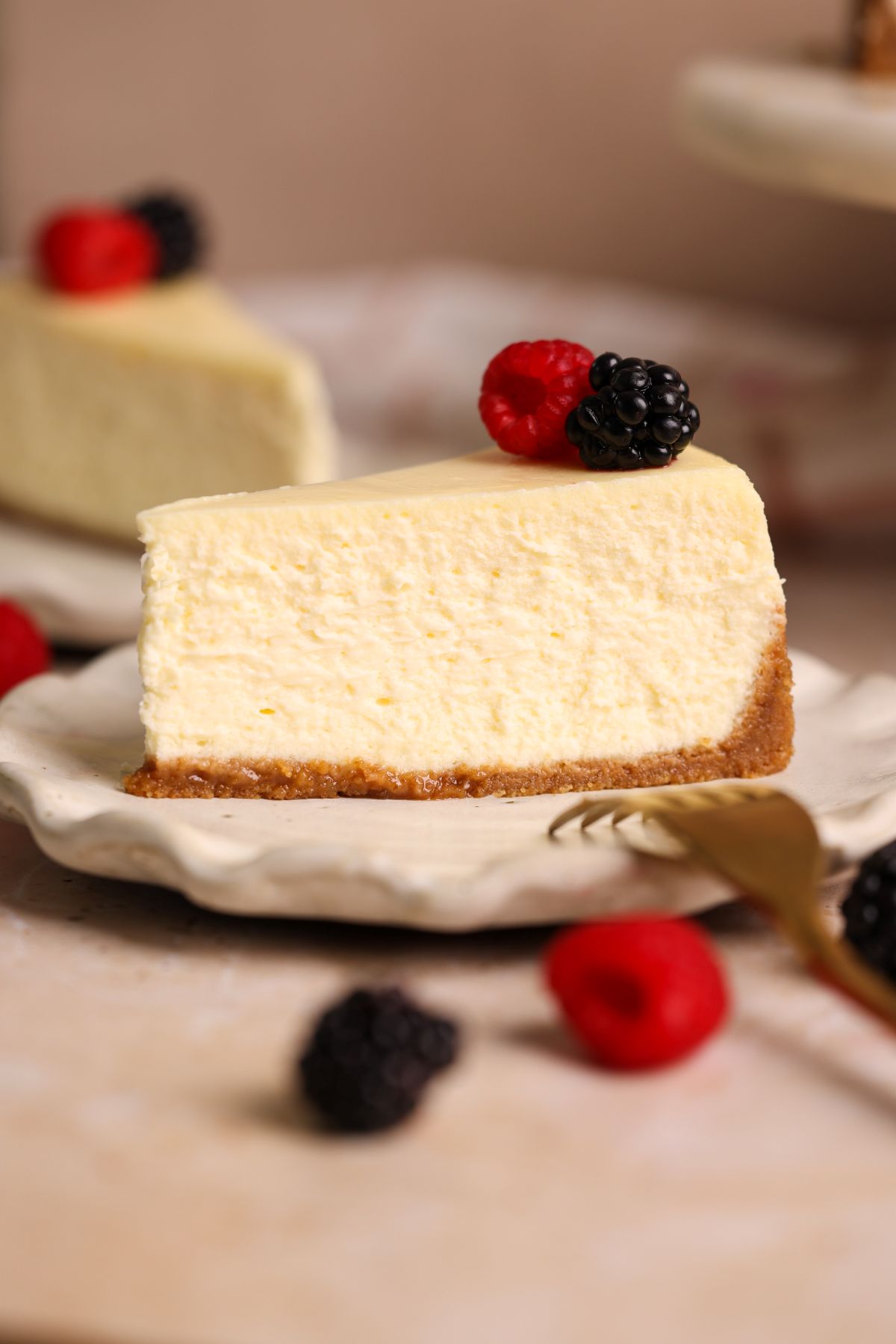 A slice of creamy cheesecake with a graham cracker crust, topped with a raspberry and a blackberry, sits on a white plate. A fork and more berries are nearby.