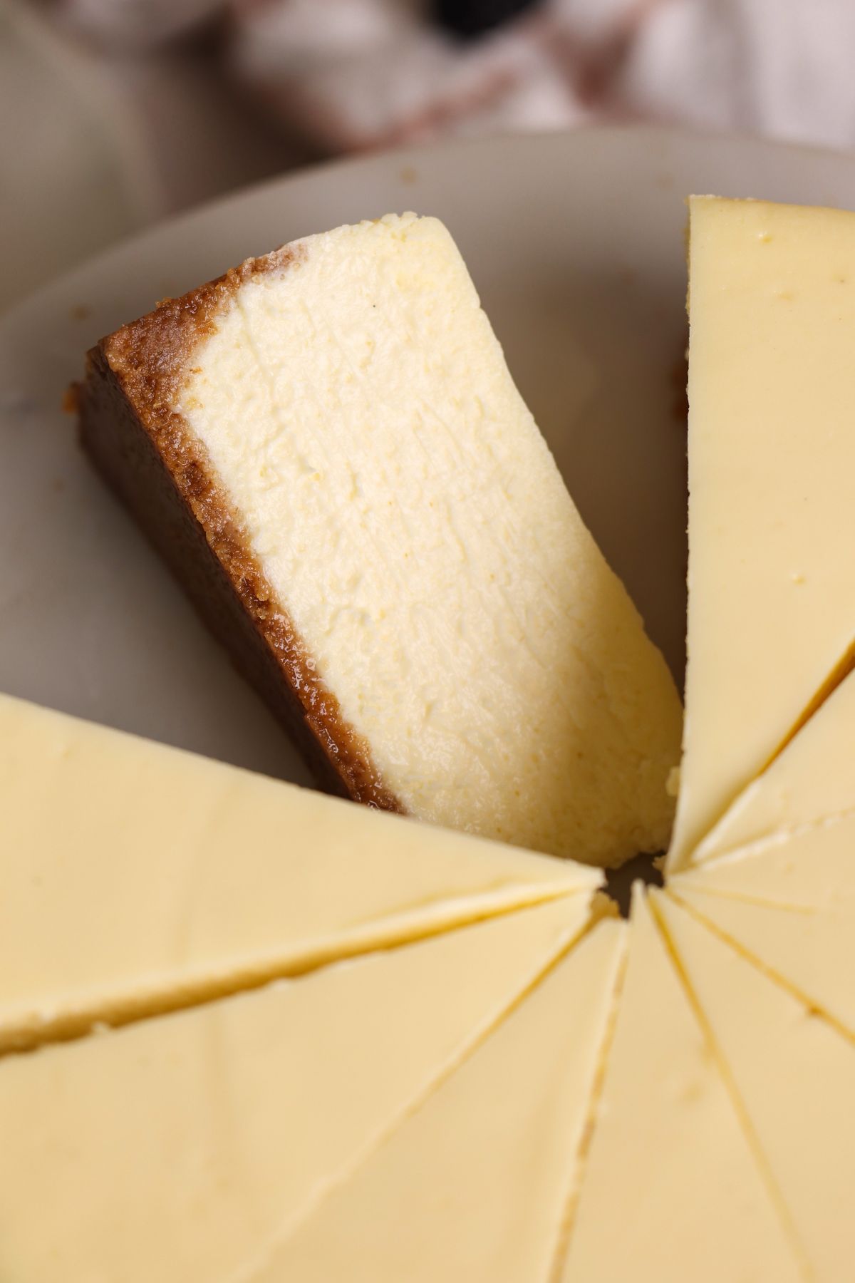 A close-up of a sliced cheesecake on a white plate, showing a thick, creamy texture and a golden-brown crust on the side of one wedge.