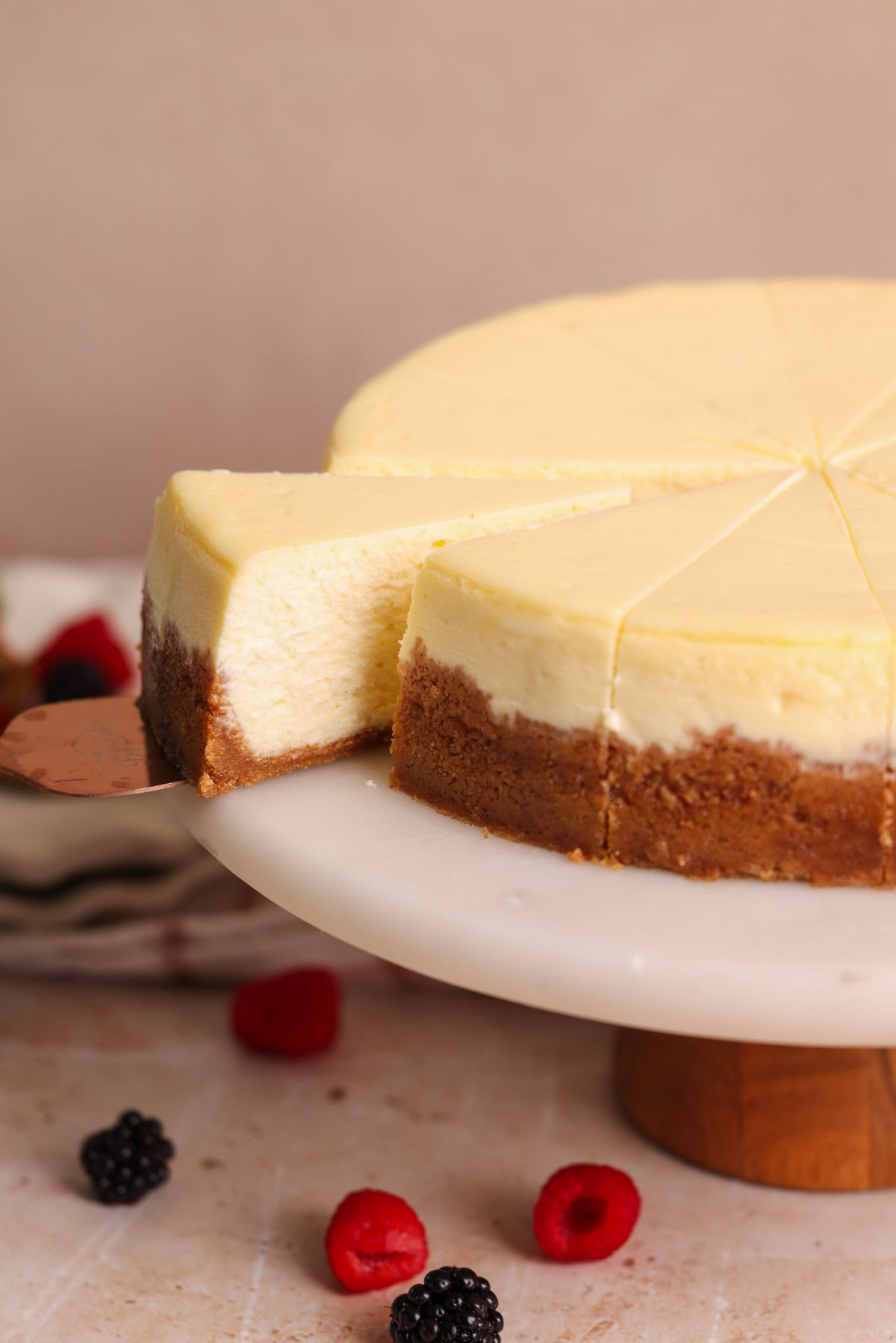 A creamy cheesecake with a graham cracker crust sits on a marble cake stand. One slice is being lifted out, and fresh berries are scattered around the base.