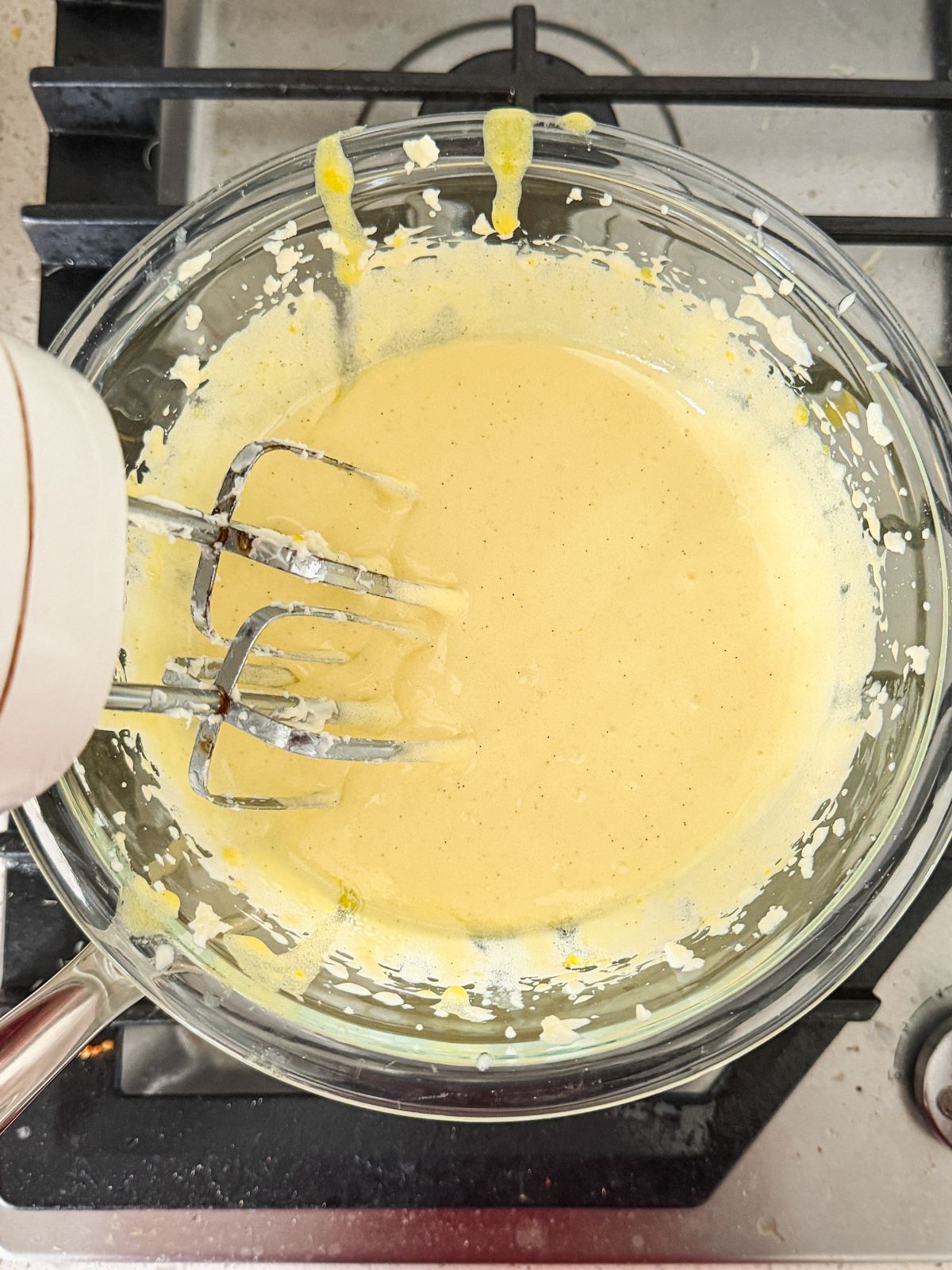 A glass bowl with whipped eggs and sugar being mixed by an electric hand mixer, sitting on a stovetop. Some batter splatters are visible on the bowls sides and on the stove.