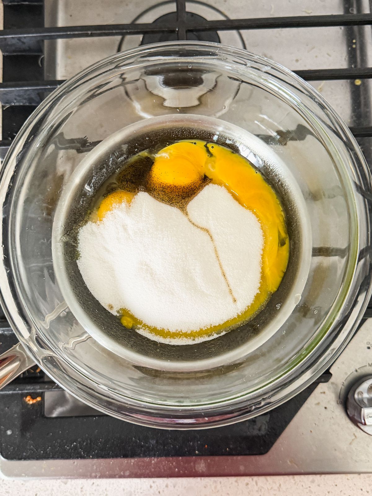 A glass bowl containing eggs and sugar sits on top of a pot over a stove burner, ready for mixing.
