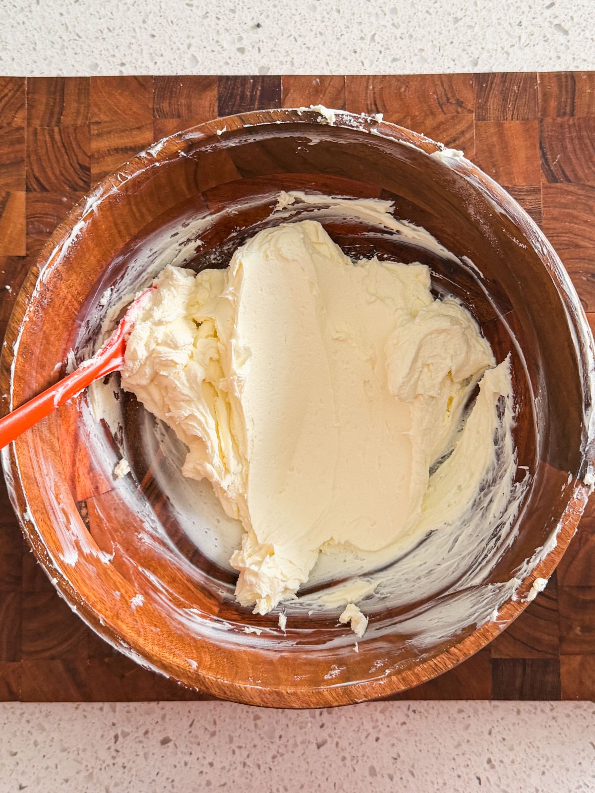 A wooden bowl with smooth, creamy mascarpone cheese being mixed by a red spatula, placed on a wooden surface.