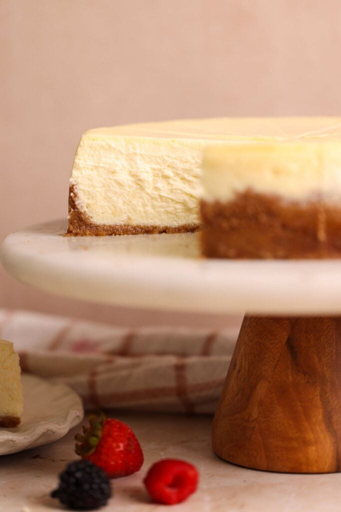 A creamy cheesecake with a graham cracker crust sits on a marble cake stand. A slice has been removed, and fresh berries are scattered on the table nearby.