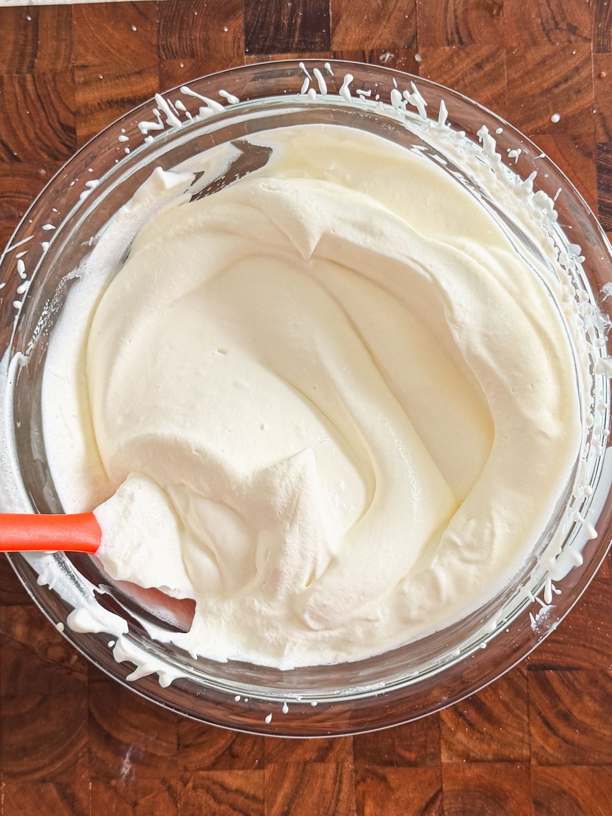 A glass bowl filled with smooth, whipped cream being mixed with a red spatula, sitting on a wooden surface.
