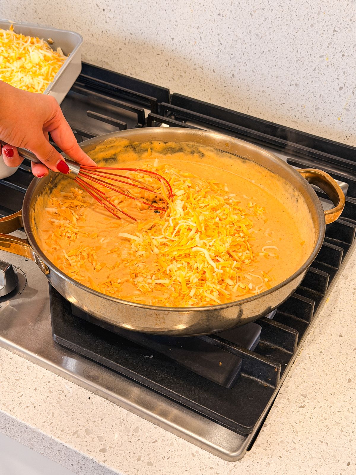 A hand holding a whisk stirs shredded cheese into a creamy sauce in a large pan on a stovetop. A baking dish with more shredded cheese sits nearby on the countertop.
