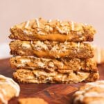 Four thick, golden biscoff cookies stacked on a wooden surface, each broken in half to reveal a gooey biscoff filling inside. Drizzles of white chocolate are visible on the cookies.