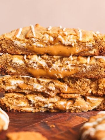 Four thick, golden biscoff cookies stacked on a wooden surface, each broken in half to reveal a gooey biscoff filling inside. Drizzles of white chocolate are visible on the cookies.