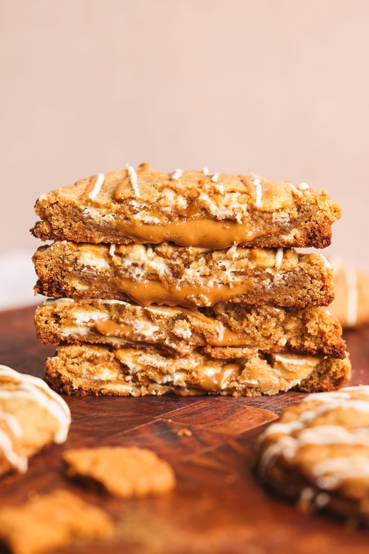 A stack of gooey biscoff cookies filled with creamy biscoff filling, topped with a light white chocolate drizzle, sits on a wooden surface with cookie crumbs nearby.