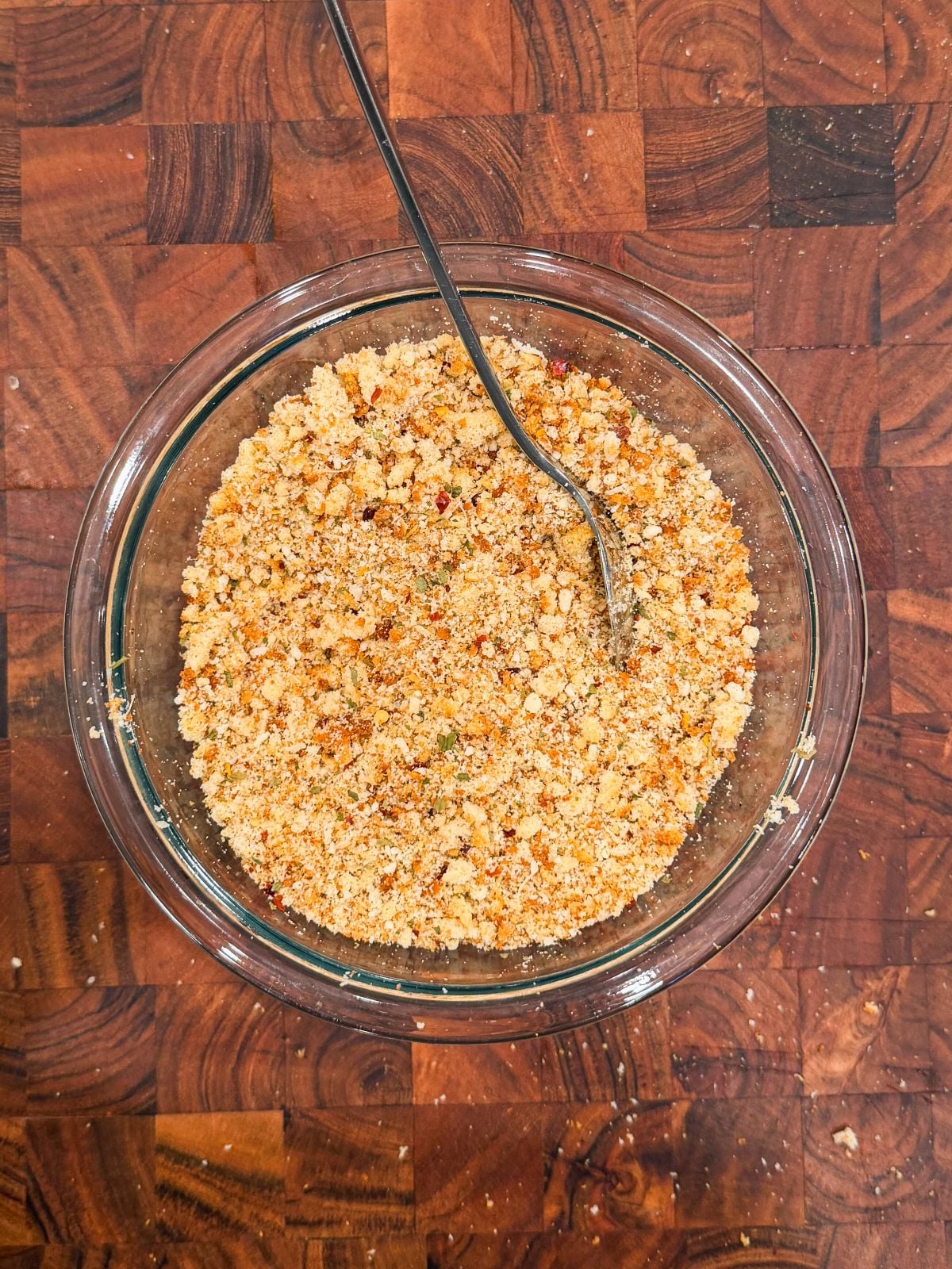 A glass bowl filled with seasoned breadcrumbs and a metal spoon, placed on a wooden cutting board.