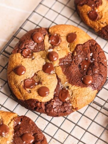 A large, round brookie cookie with brownie and cookie dough swirled together, topped with chocolate chips and a sprinkle of sea salt, cooling on a wire rack.