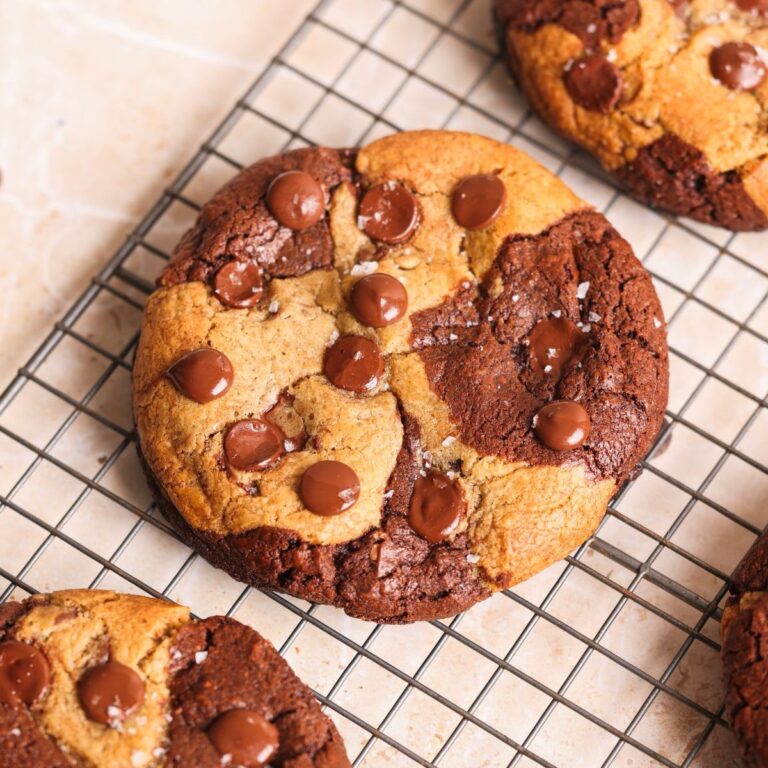 A large, round brookie cookie with brownie and cookie dough swirled together, topped with chocolate chips and a sprinkle of sea salt, cooling on a wire rack.