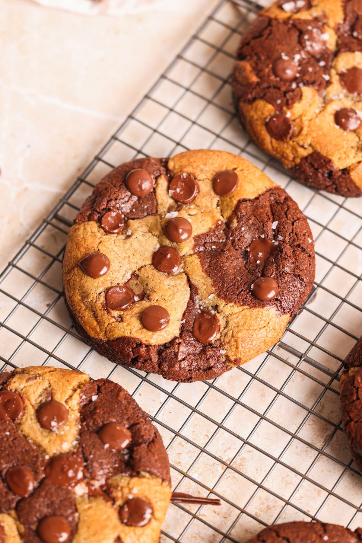 A large, round brookie cookie with swirls of brownie and cookie dough, topped with melted chocolate chips, sits on a wire cooling rack with other similar cookies nearby.