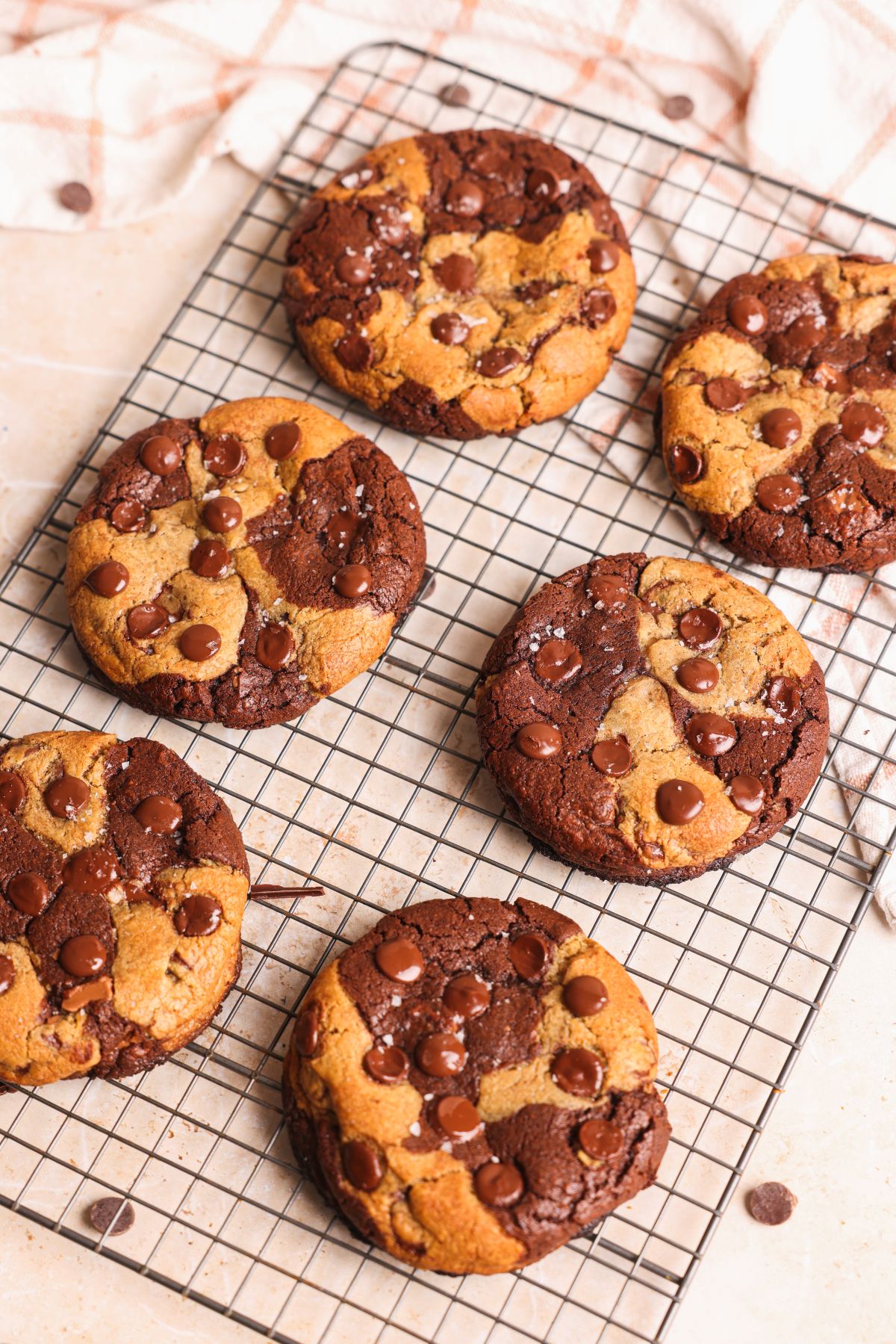Six large brookie cookies with swirls of brownie and cookie dough, packed with chocolate chips, cool on a wire rack. A light cloth with a grid pattern is partially visible in the background.
