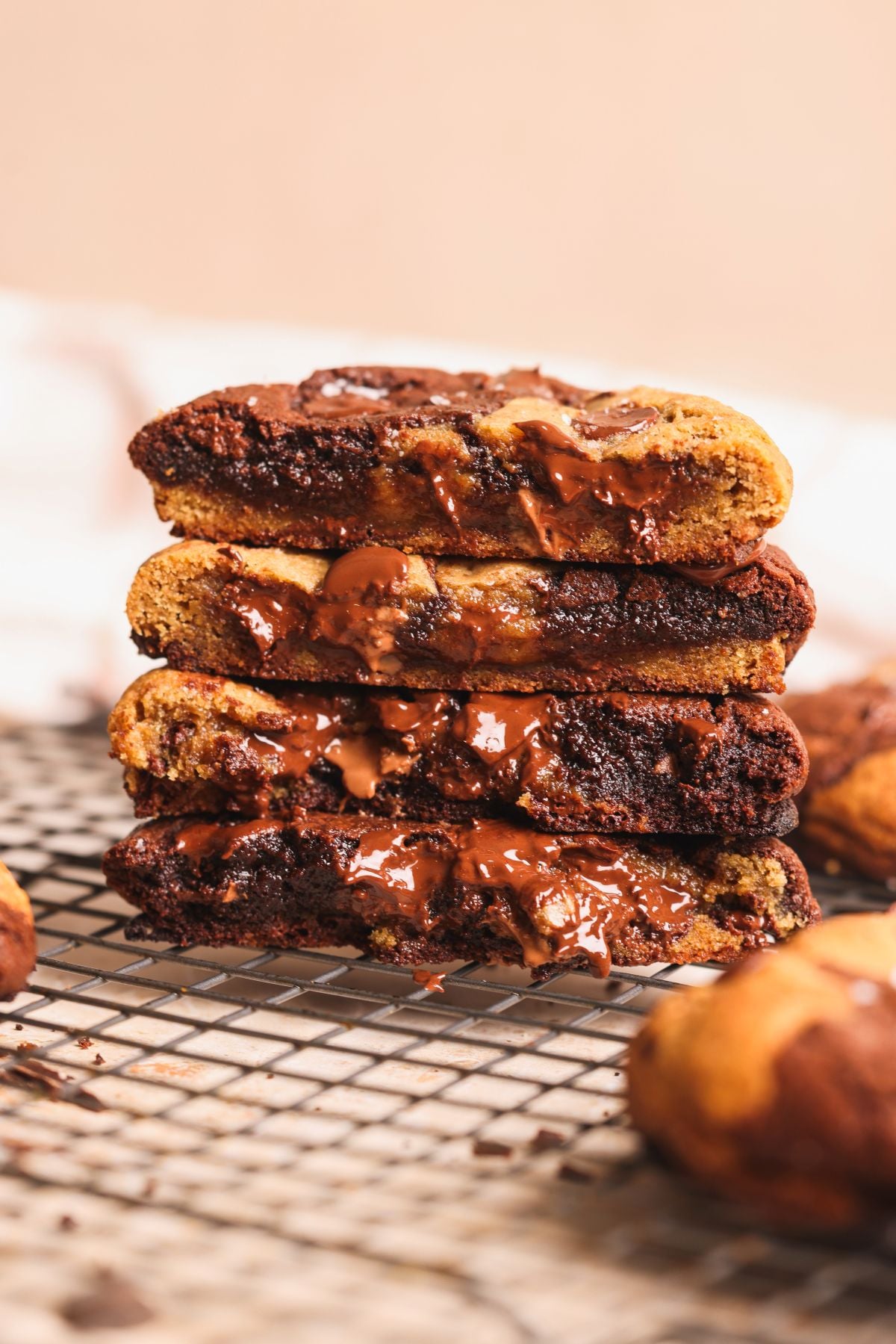 A stack of gooey brookie cookies, broken in half to reveal layers of melted chocolate and cookie dough inside, sits on a cooling rack with a blurred background.