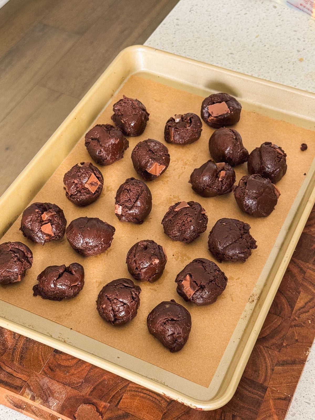 A baking tray lined with parchment paper holds twenty scoops of brownie cookie dough, some containing visible chocolate chunks. The tray sits on a wooden cutting board and a kitchen counter.