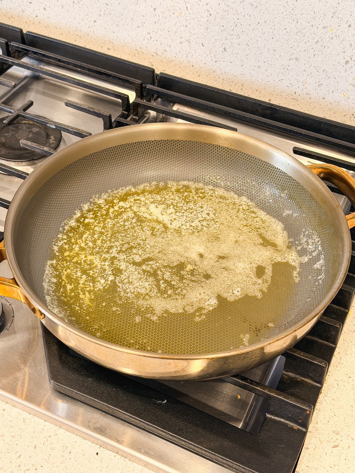 A frying pan with melted butter sits on a gas stove, with small bubbles forming in the butter as it heats. The stove and speckled countertop are visible in the background.