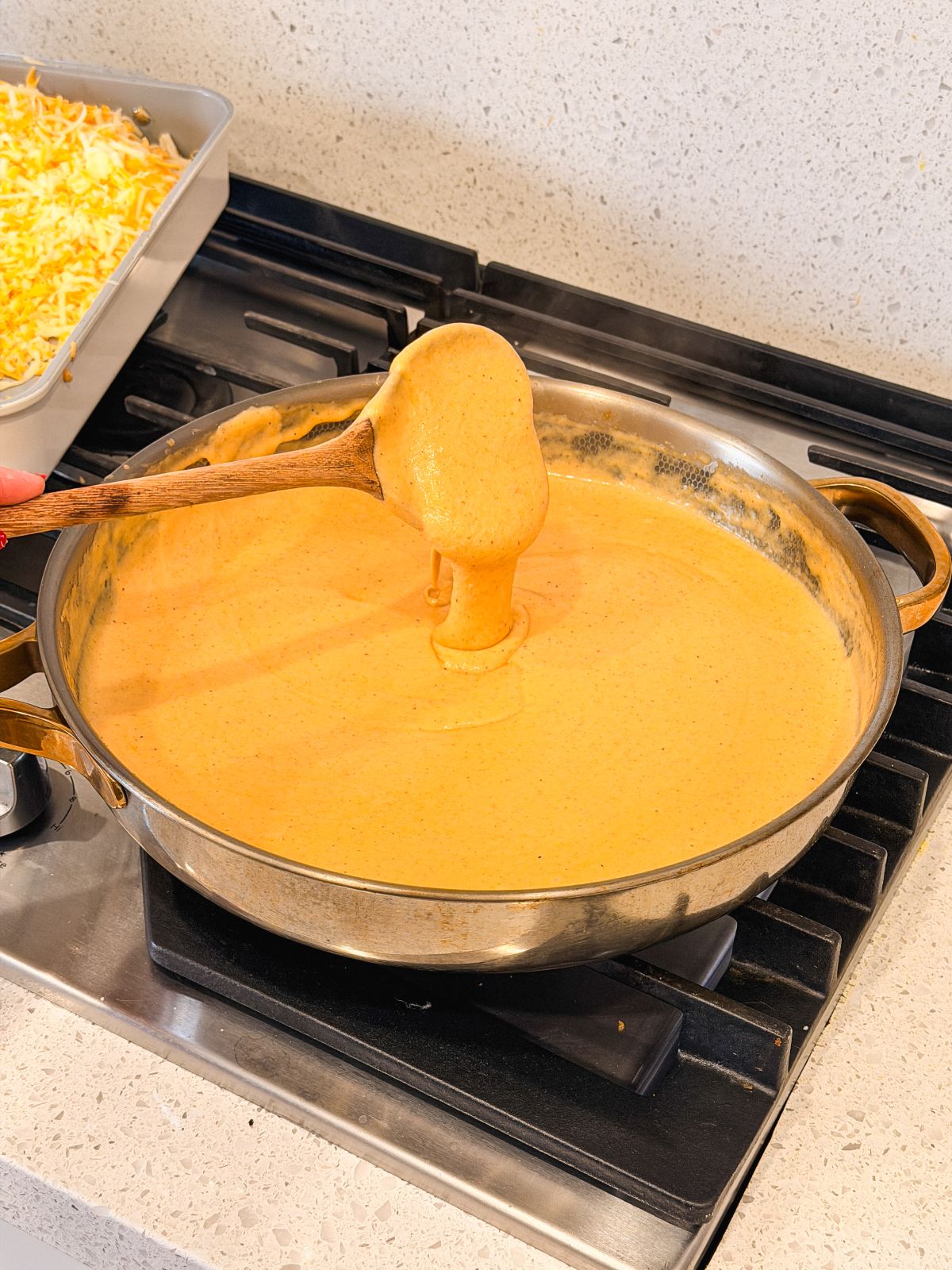 A wooden spoon lifts creamy, orange cheese sauce from a large skillet on a stove, with a tray of shredded cheese visible nearby on a white countertop.