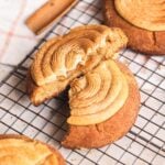 A soft churro cookie with a swirl of caramel-colored dulce de leche cream cheese frosting is broken in half on a cooling rack, revealing a gooey dulce de leche filling inside. Other cookies and a cinnamon stick are visible nearby.