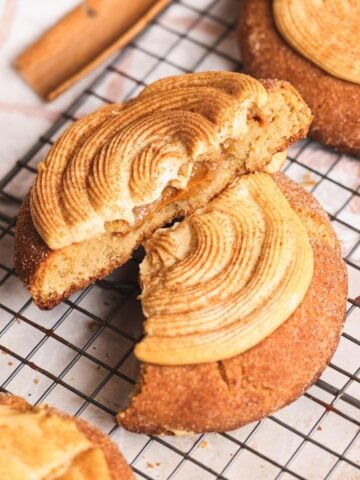 A soft churro cookie with a swirl of caramel-colored dulce de leche cream cheese frosting is broken in half on a cooling rack, revealing a gooey dulce de leche filling inside. Other cookies and a cinnamon stick are visible nearby.