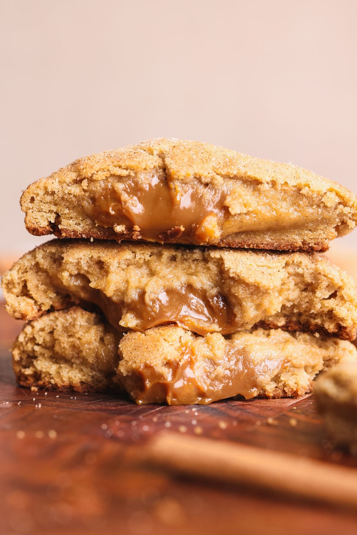 Three thick churro cookies stacked, each broken in half to reveal gooey dulce de leche filling oozing from the center. The cookies have a golden-brown, gooey texture and sit on a wooden surface.