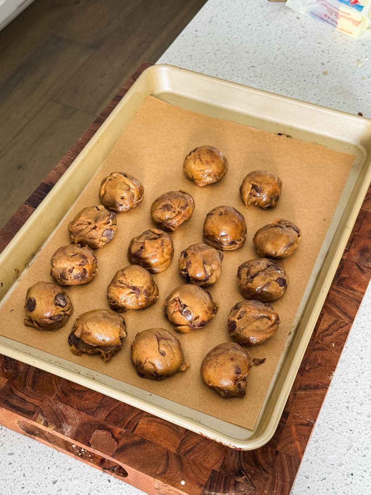 A baking tray lined with parchment paper holds 16 balls of raw chocolate chip cookie dough. The tray rests on a wooden cutting board atop a kitchen counter.