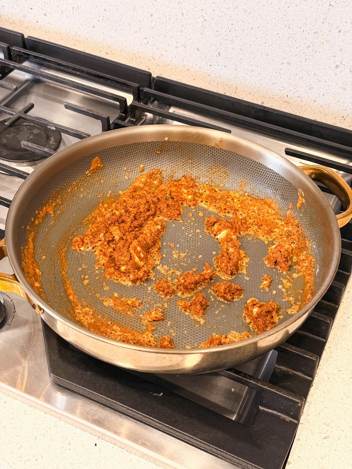 A large metal frying pan on a stovetop contains a partially cooked, reddish-brown roux base with cream cheese. The mixture is spread unevenly, with some sticking to the bottom of the pan. The stove and countertop are visible.