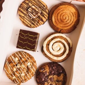 Assorted cookies and a brownie in a box, including swirled, drizzled, and chocolate chip varieties, arranged neatly on a light surface.