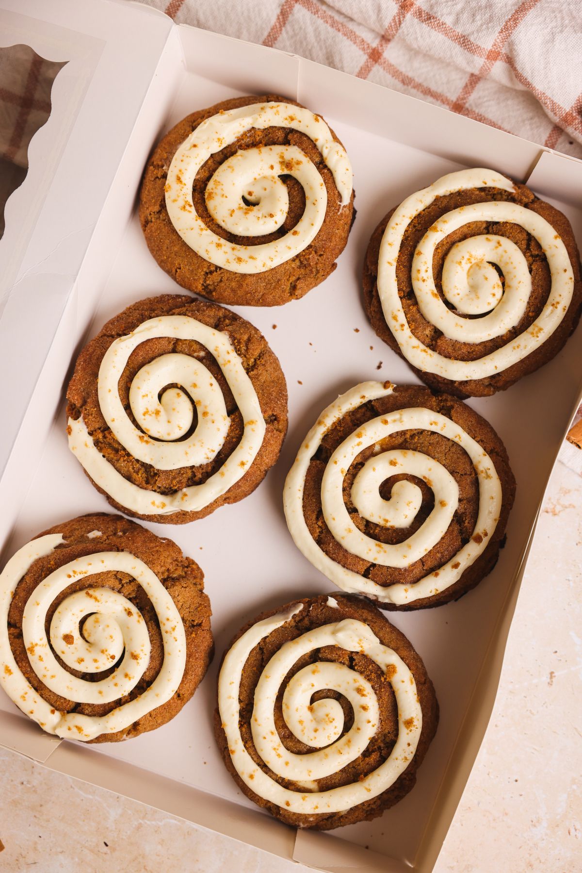 A box of six large pumpkin cinnamon roll cookies, each topped with cream cheese icing swirled in a spiral pattern, arranged neatly inside a white bakery box on a light surface.