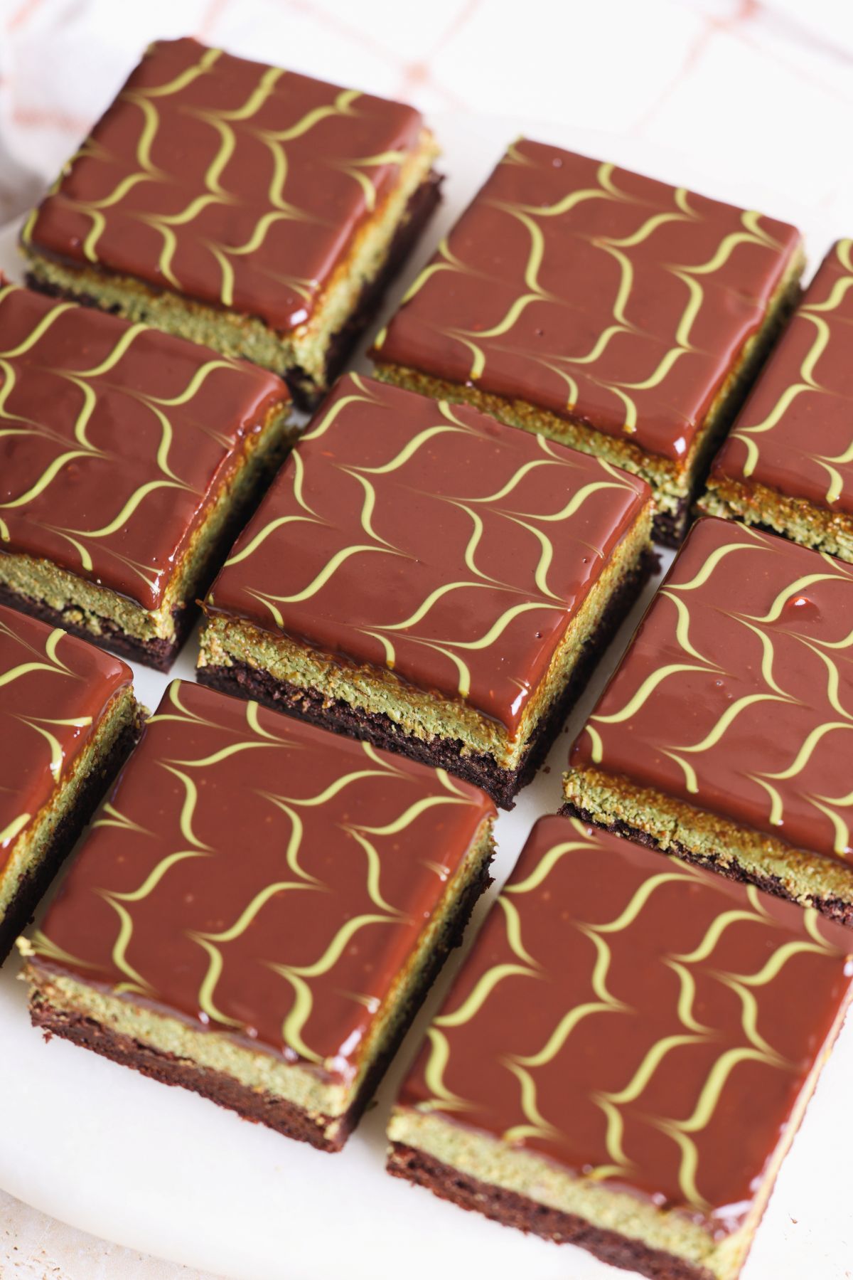 Nine Dubai chocolate brownies, feature a glossy ganache glaze with a green pistachio drizzle. Arranged neatly on a white surface, each bar reveals distinct pistachio and brownie layers beneath the rich chocolate topping.