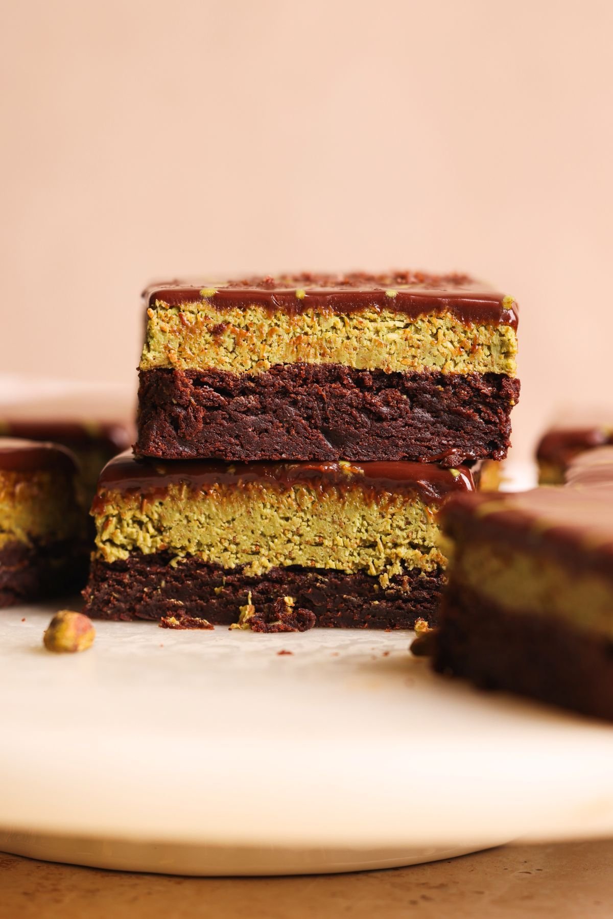 Two stacked dubai chocolate brownies with a thick chocolate brownie base, a green pistachio knafeh layer in the middle, and a glossy ganache topping, displayed on a light-colored surface.