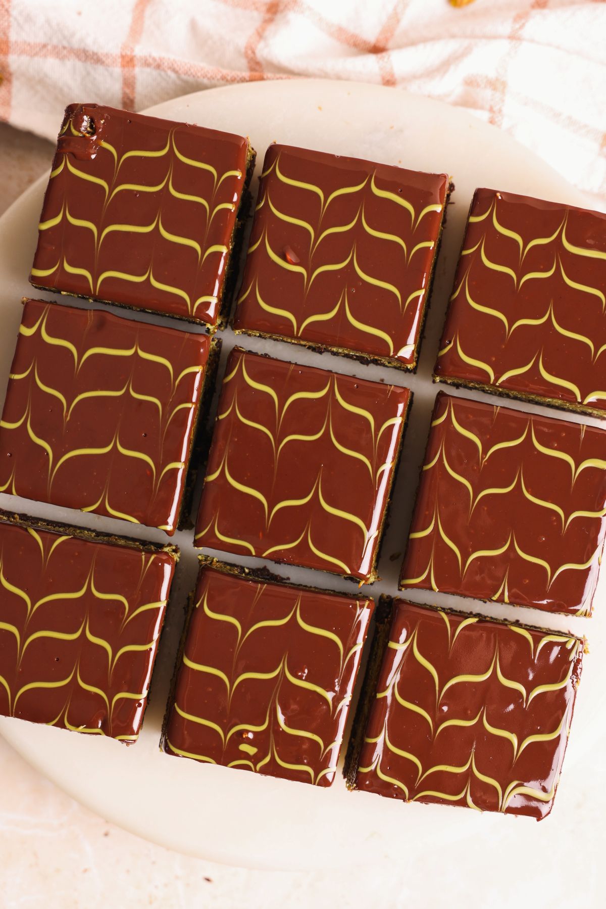 Nine dubai chocolate brownies topped with a rich ganache are arranged closely on a white plate. The chocolate topping is decorated with a green pistachio feathered pattern, creating a marbled effect on each pastry.