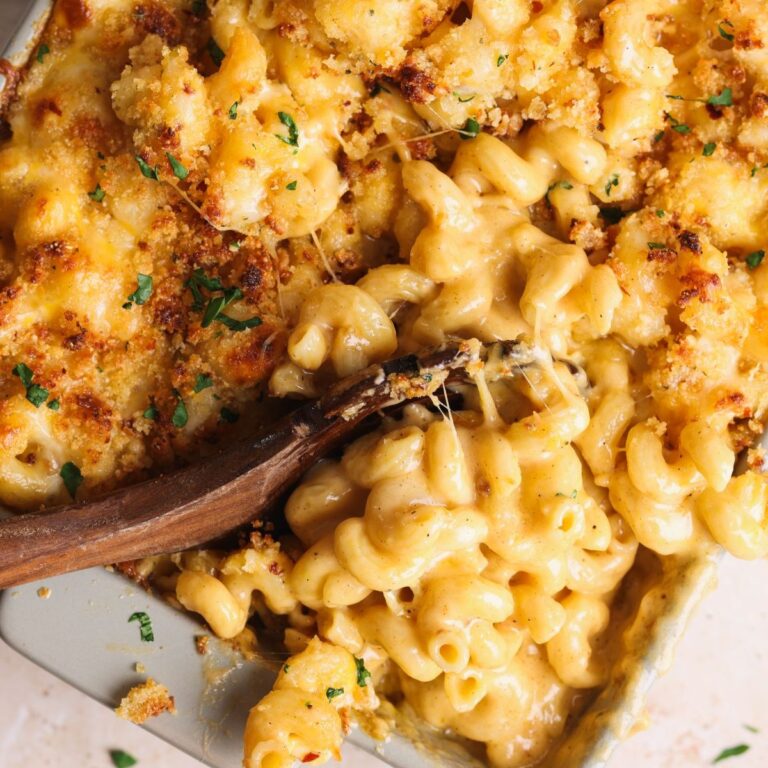 A close-up of creamy baked macaroni and cheese with a golden, crispy breadcrumb topping, being scooped from a baking dish with a wooden spoon.