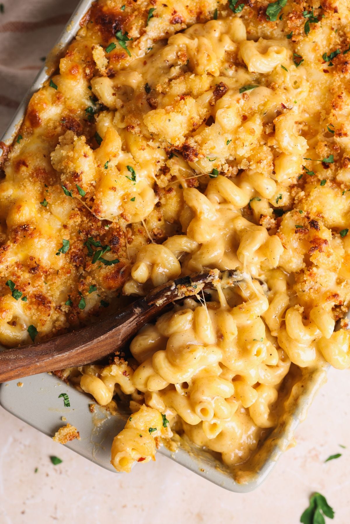 Close-up of creamy baked macaroni and cheese topped with golden, crispy breadcrumbs and garnished with chopped parsley. A wooden spoon is scooping out a portion from the casserole dish.