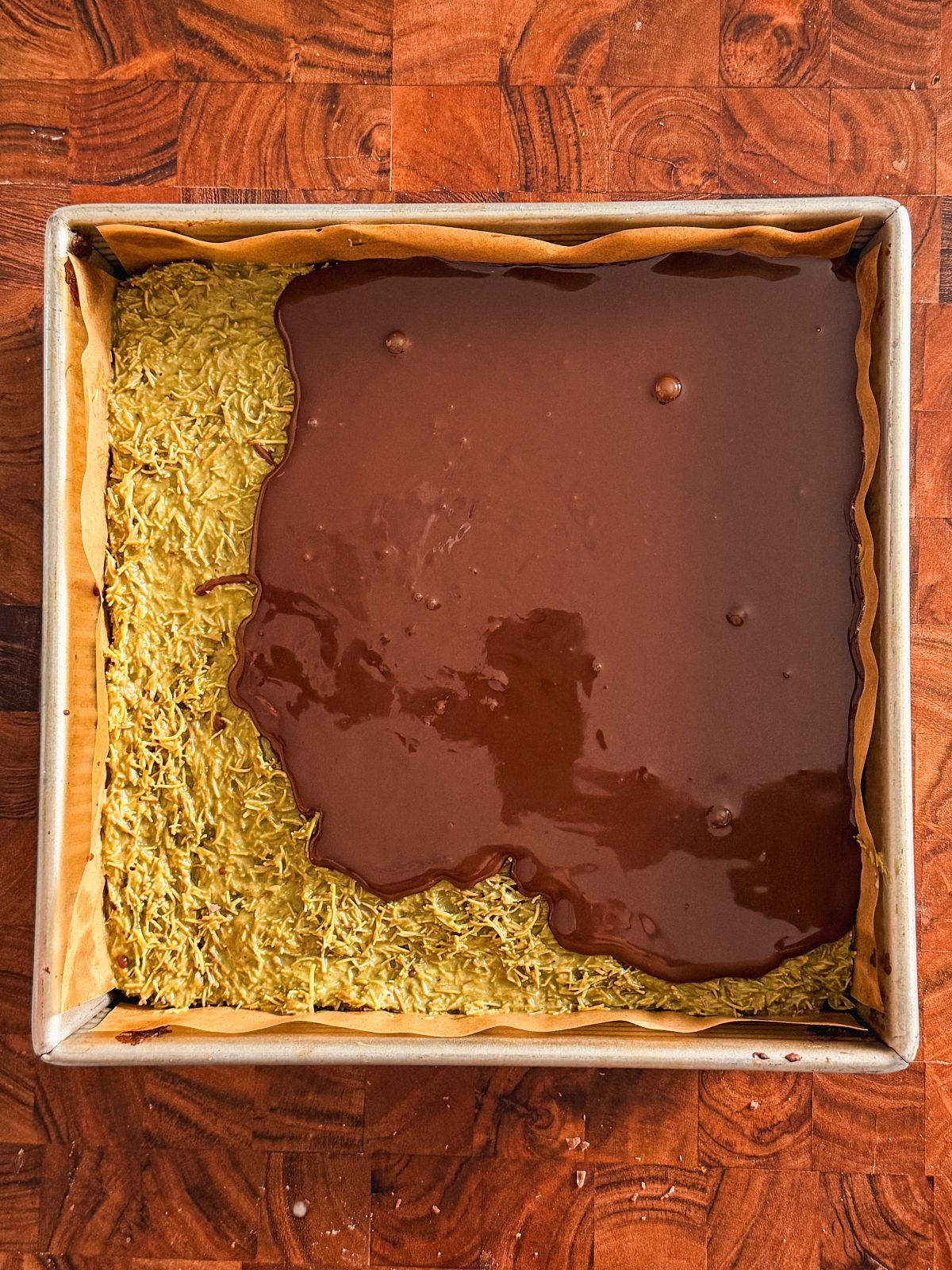 A baking tray lined with parchment paper contains a shredded, pistachio knafeh layer with ganache being poured over about half of it, sitting on a wooden surface.