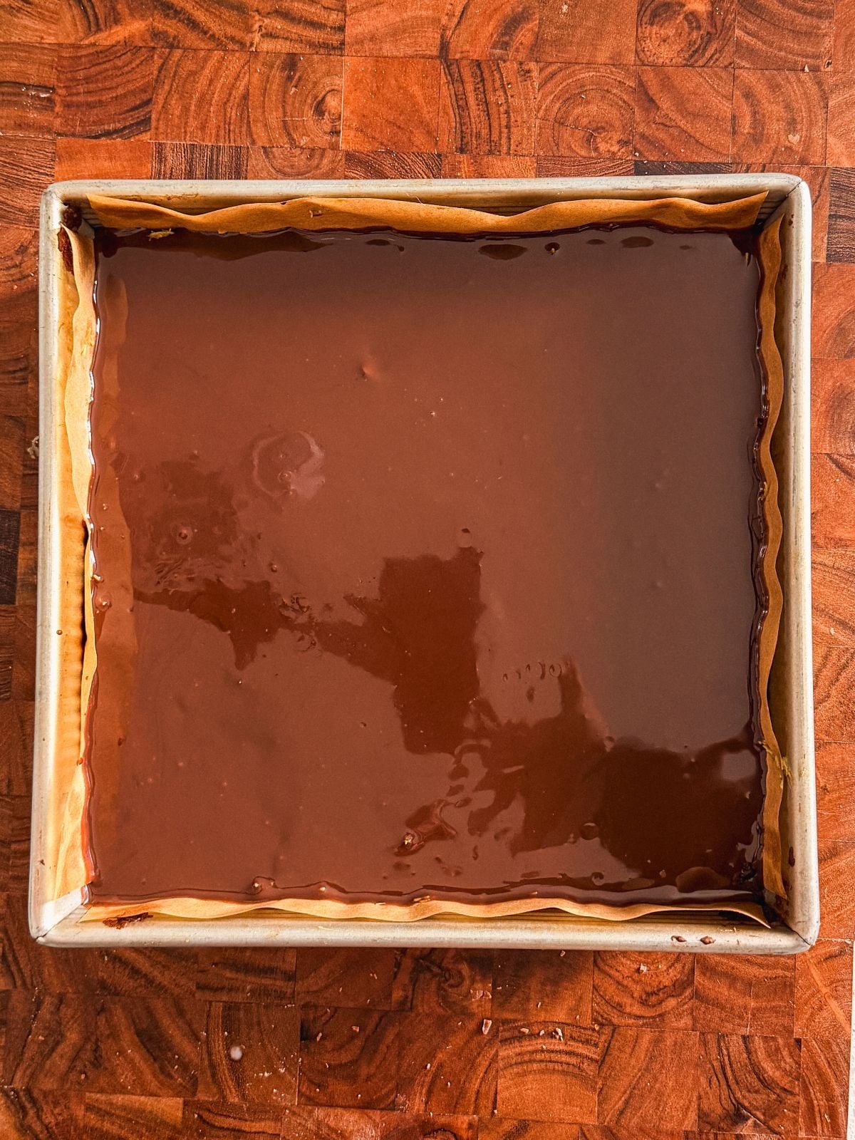 A square baking pan lined with parchment paper holds dubai chocolate brownies with a smooth layer of glossy ganache, resting on a wooden surface.