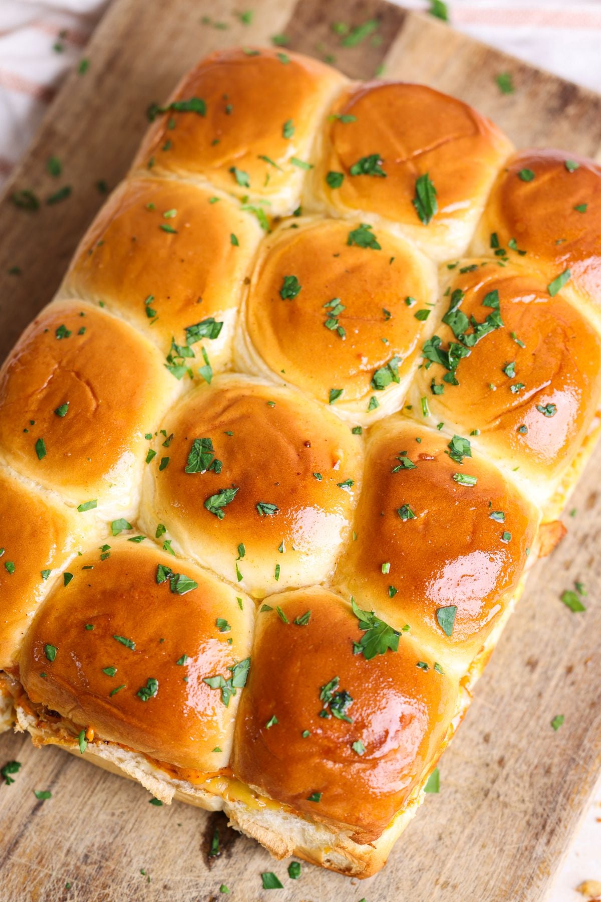 A batch of golden-brown, shiny sliders topped with honey butter and chopped parsley sits on a wooden cutting board.