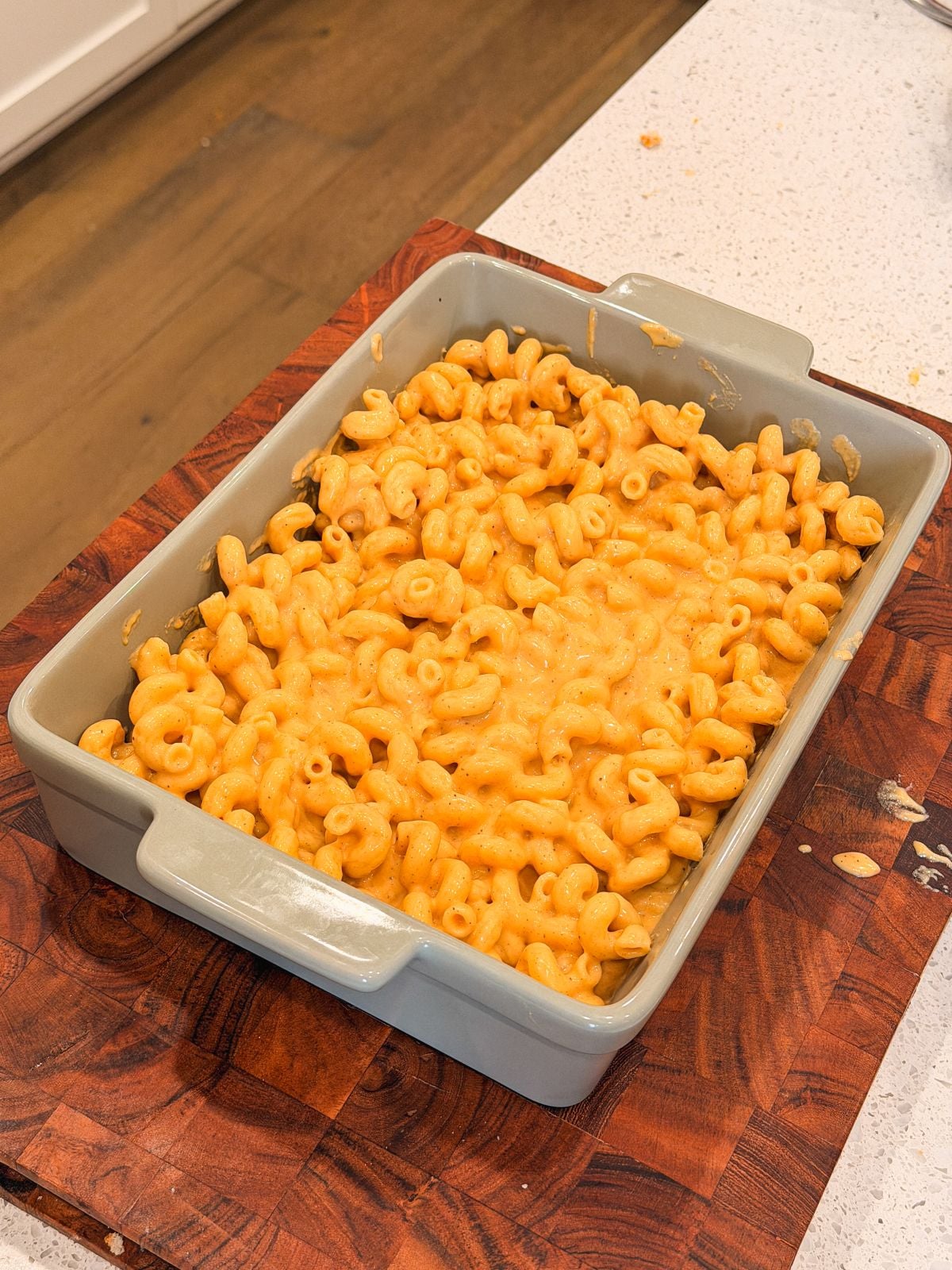 A rectangular gray baking dish half filled with creamy macaroni and cheese sits on a wooden cutting board on a white countertop.
