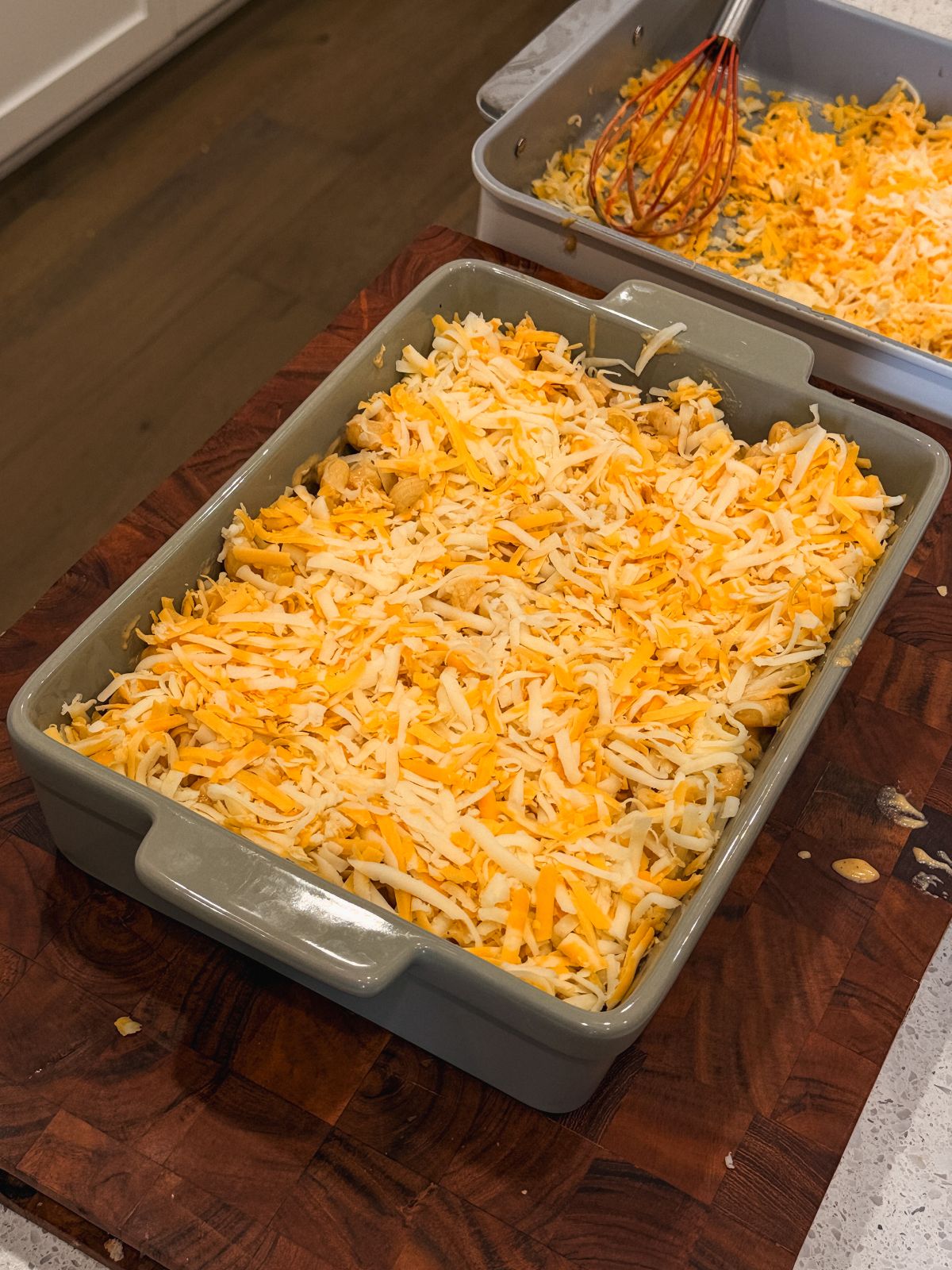 A rectangular baking dish filled with macaroni and cheese, topped with shredded yellow and white cheese, sits on a wooden cutting board. In the background, another pan and a red whisk are visible.