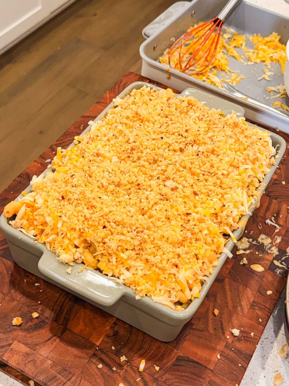 A baking dish filled with macaroni and cheese topped with grated cheese and breadcrumbs sits on a wooden cutting board, with a whisk and baking tray in the background.