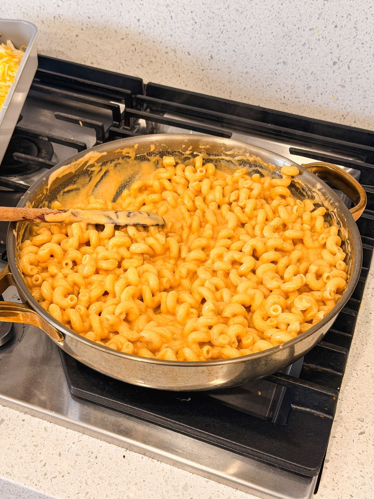 A skillet filled with creamy macaroni and cheese sits on a stovetop, with a wooden spoon resting in the pasta. The cheese sauce coats the macaroni evenly. The stove and countertop appear clean and tidy.