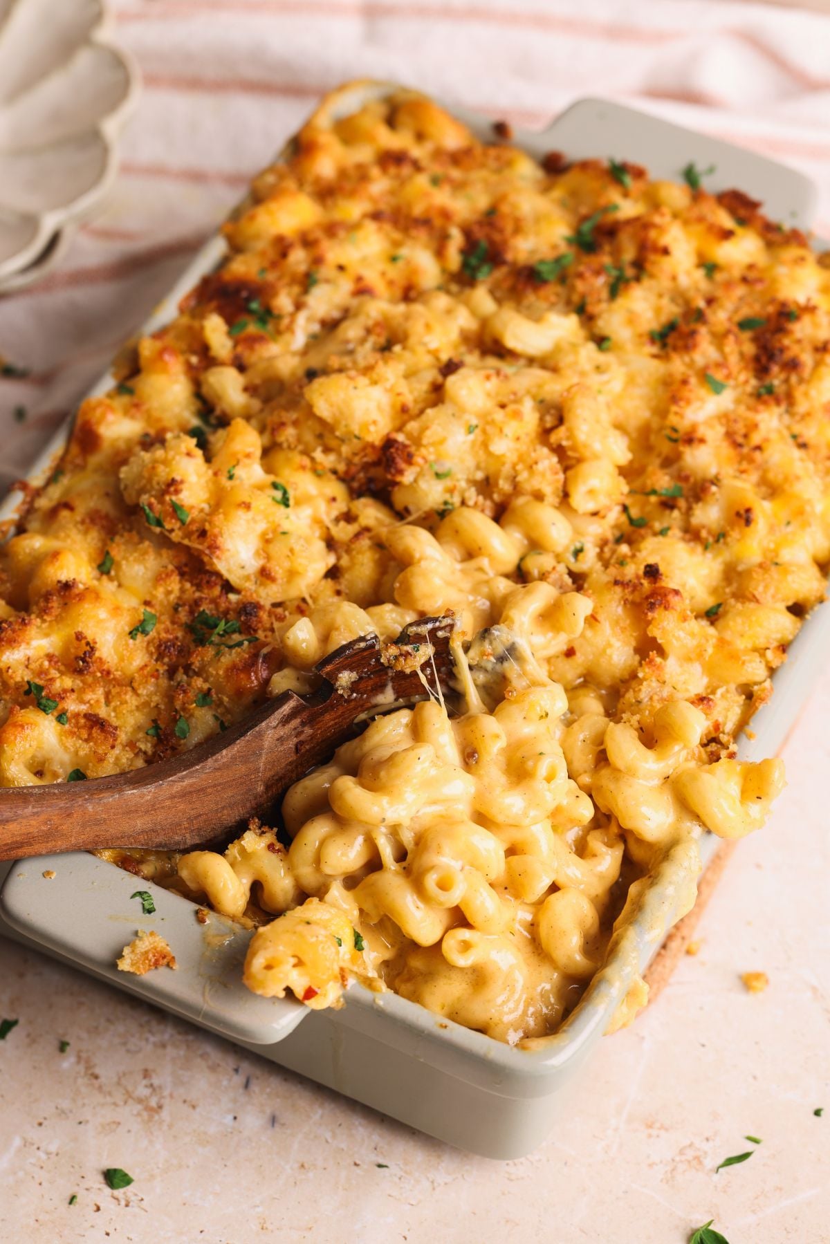 A baking dish filled with creamy, baked macaroni and cheese topped with golden, crispy breadcrumbs. A wooden spoon is scooping out a serving, showing the melted cheese and spiral pasta inside.